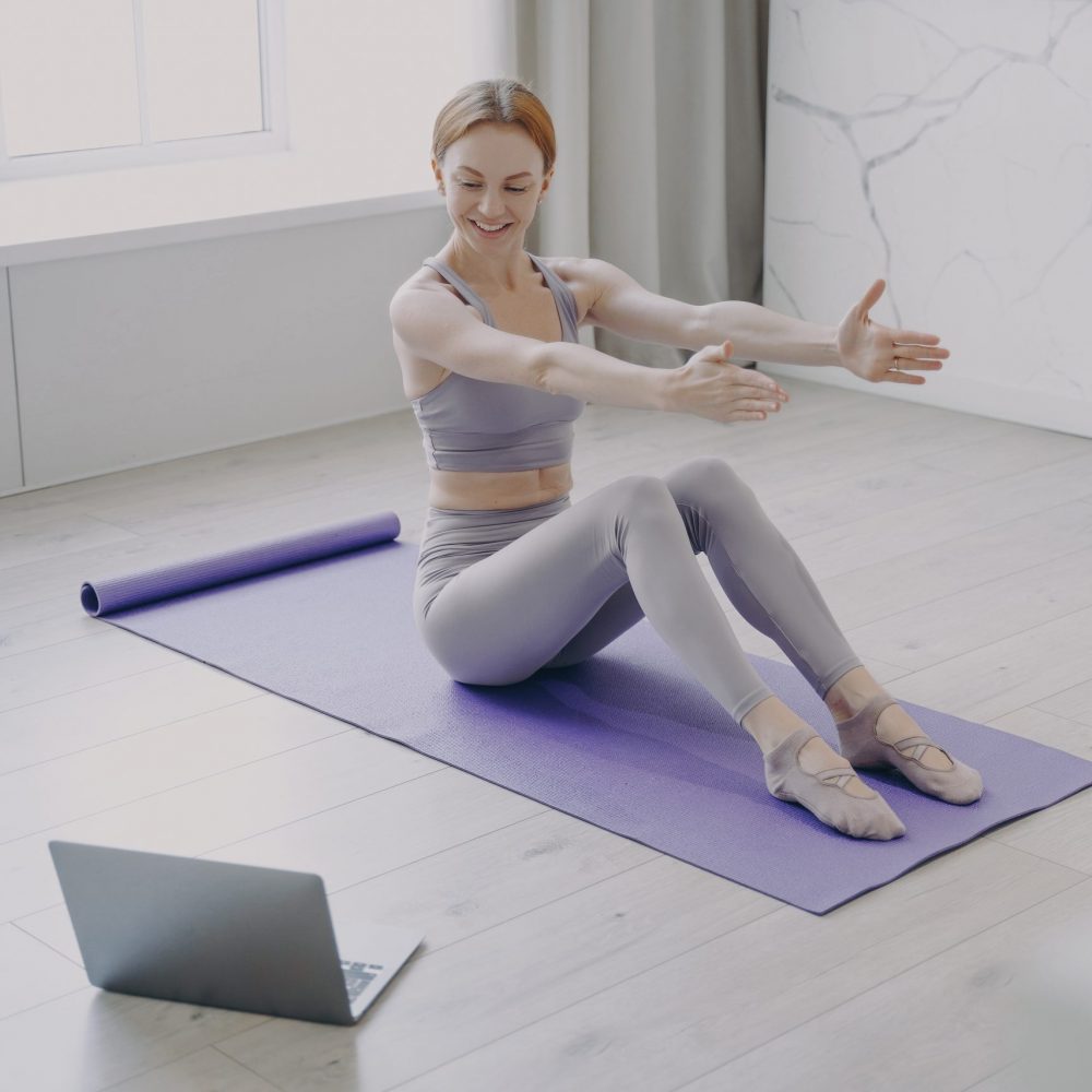 Slim sporty caucasian girl in sportswear is stretching on mat at home. Active healthy lifestyle. Concept of distance learning and online fitness classes. Personal training on coronavirus quarantine.